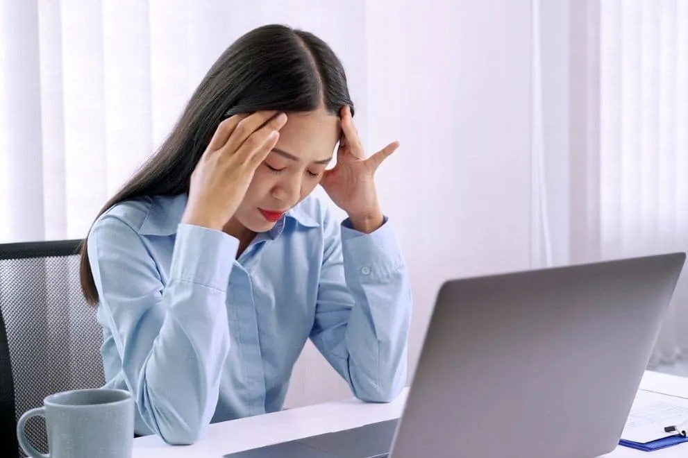 Stressed woman sitting at a desk with a laptop, holding her head in discomfort.