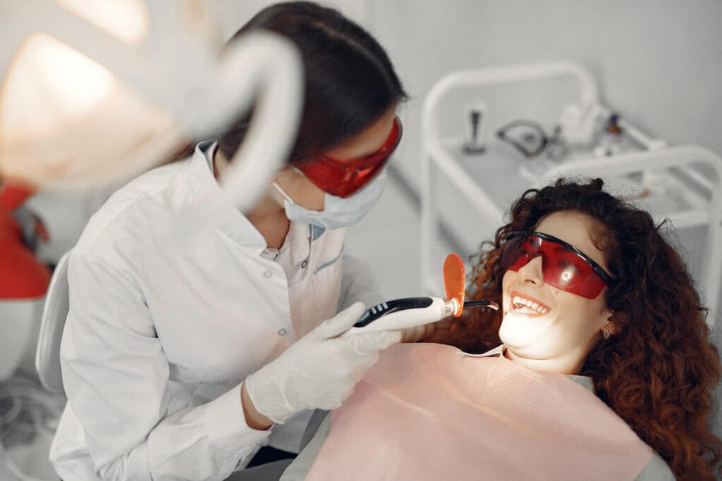 Dentist in protective mask and eyewear working on a smiling female patient wearing red protective glasses in a modern dental office.