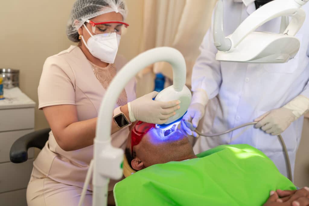 Dentist and assistant performing a procedure with blue light on a patient in a dental clinic, both wearing protective gear.