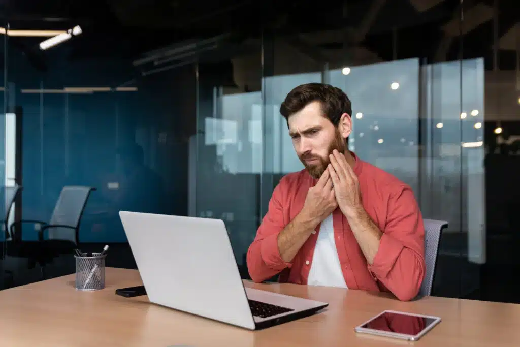 Man touching jaw while working on laptop.